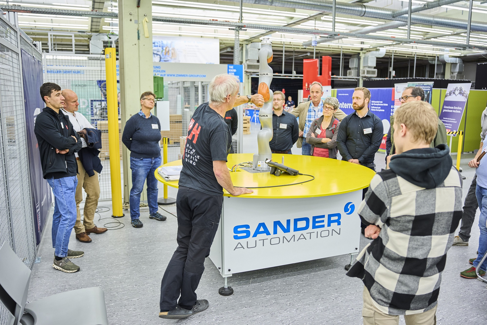 Menschen stehen um einen runden Tisch auf dem sich ein Roboterarm befindet. Ein Mann erklärt etwas