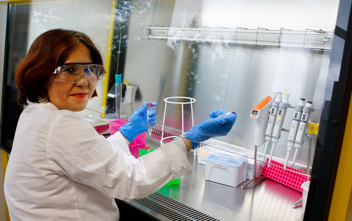 Eine Frau im weißen Kittel mit Laborbrille und Handschuhe sitzt in einem Labor und arbeitet mit Pipette und Reagenzglas