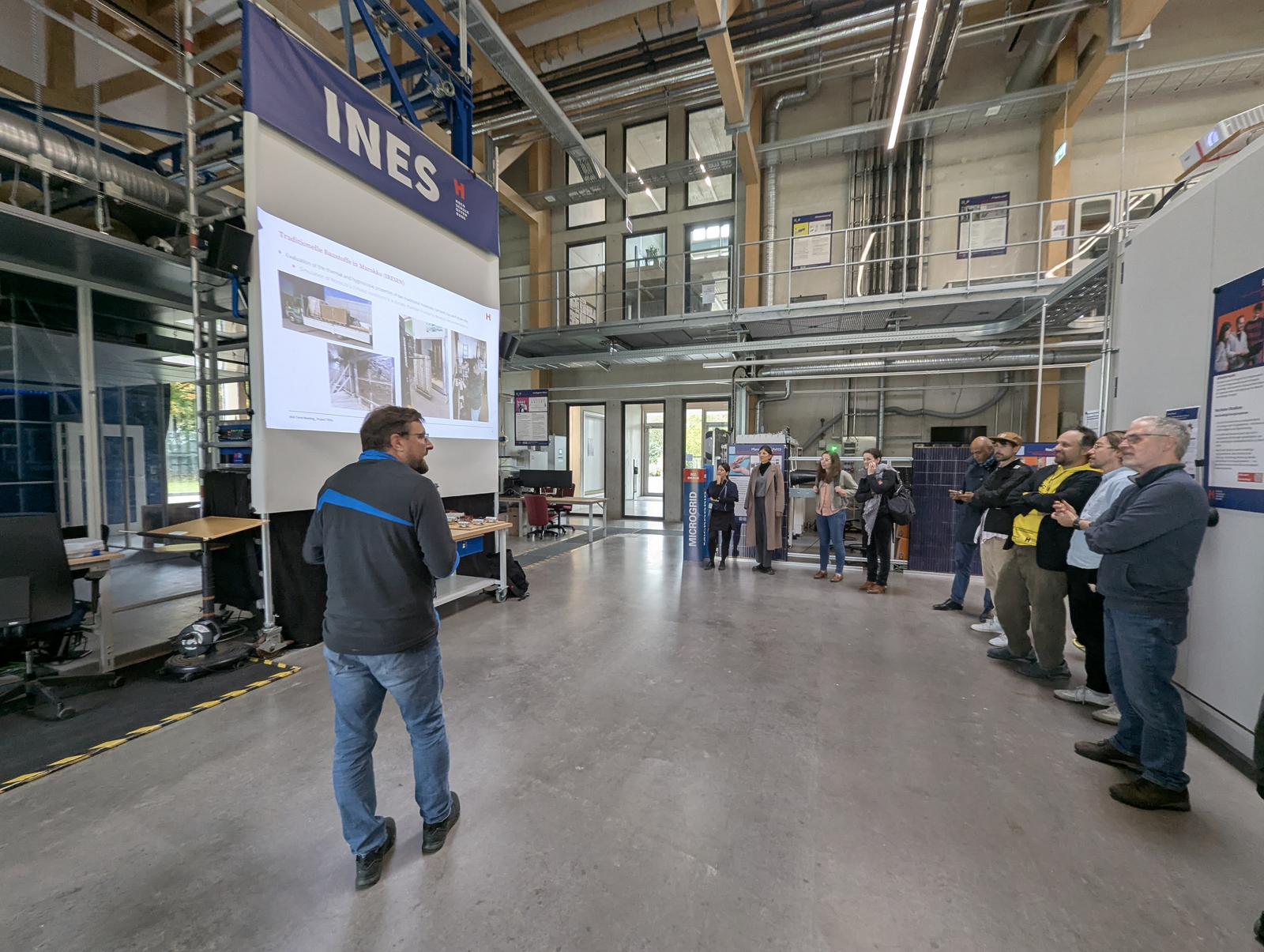 2025-10-24 Web-Meldung TRAIL Midterm Meeting Rundgang Personen stehen in einem Technikum vor einer Leinwand auf der eine Präsentation zu sehen ist