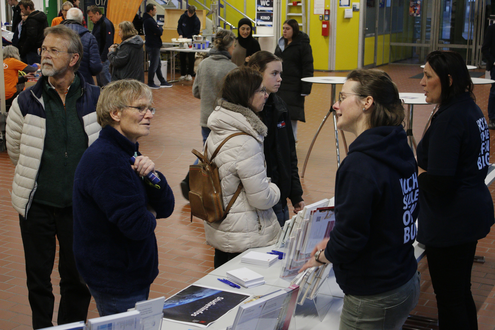 Zwei junge Frauen in Hochschulhoodies sprechen am Studieninfotisch mit Besucherinnen und Besuchern