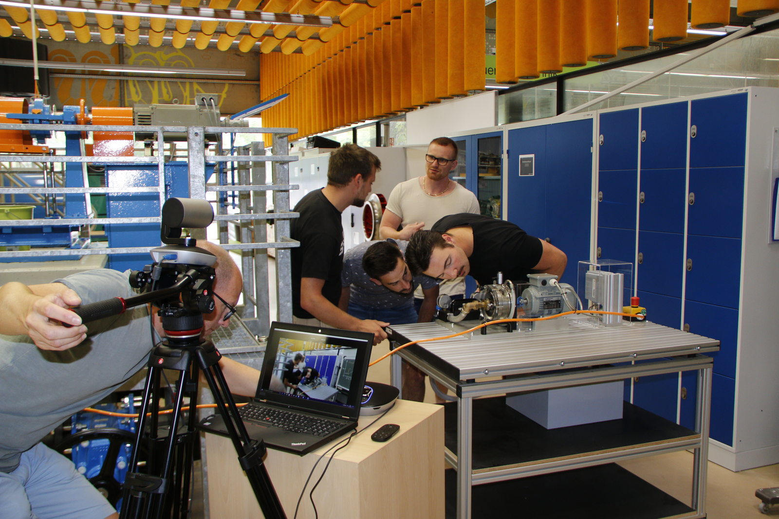 Studierende beugen sich neugierig über eine Maschine und werden dabei von einem KAmermann gefilmt